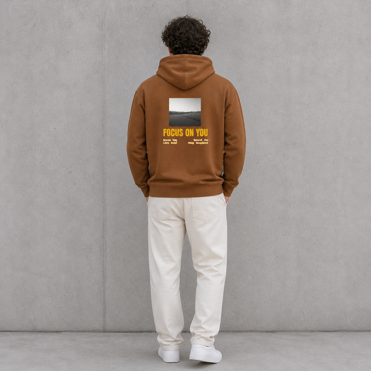 Person wearing a brown hoodie with 'Focus on You' text on the back against a gray wall.