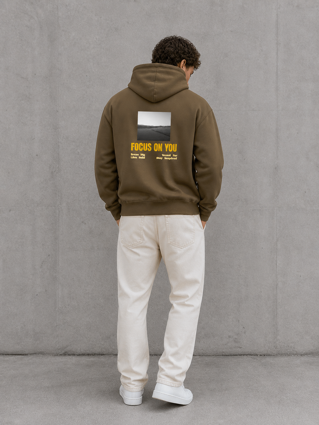 Person wearing a brown hoodie with a graphic and text on the back, standing against a gray wall.