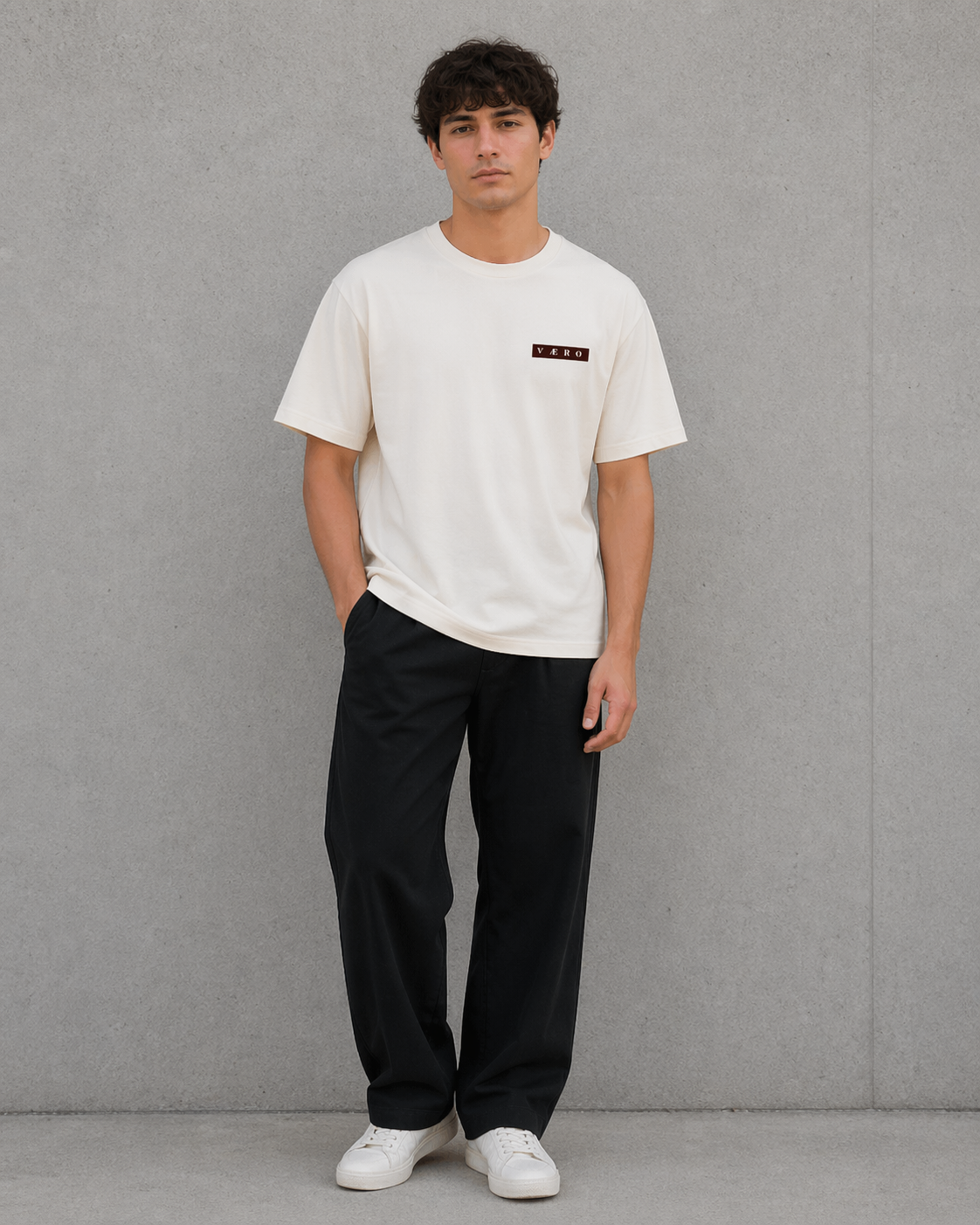 Man wearing a white t-shirt with a small logo and black pants against a gray background