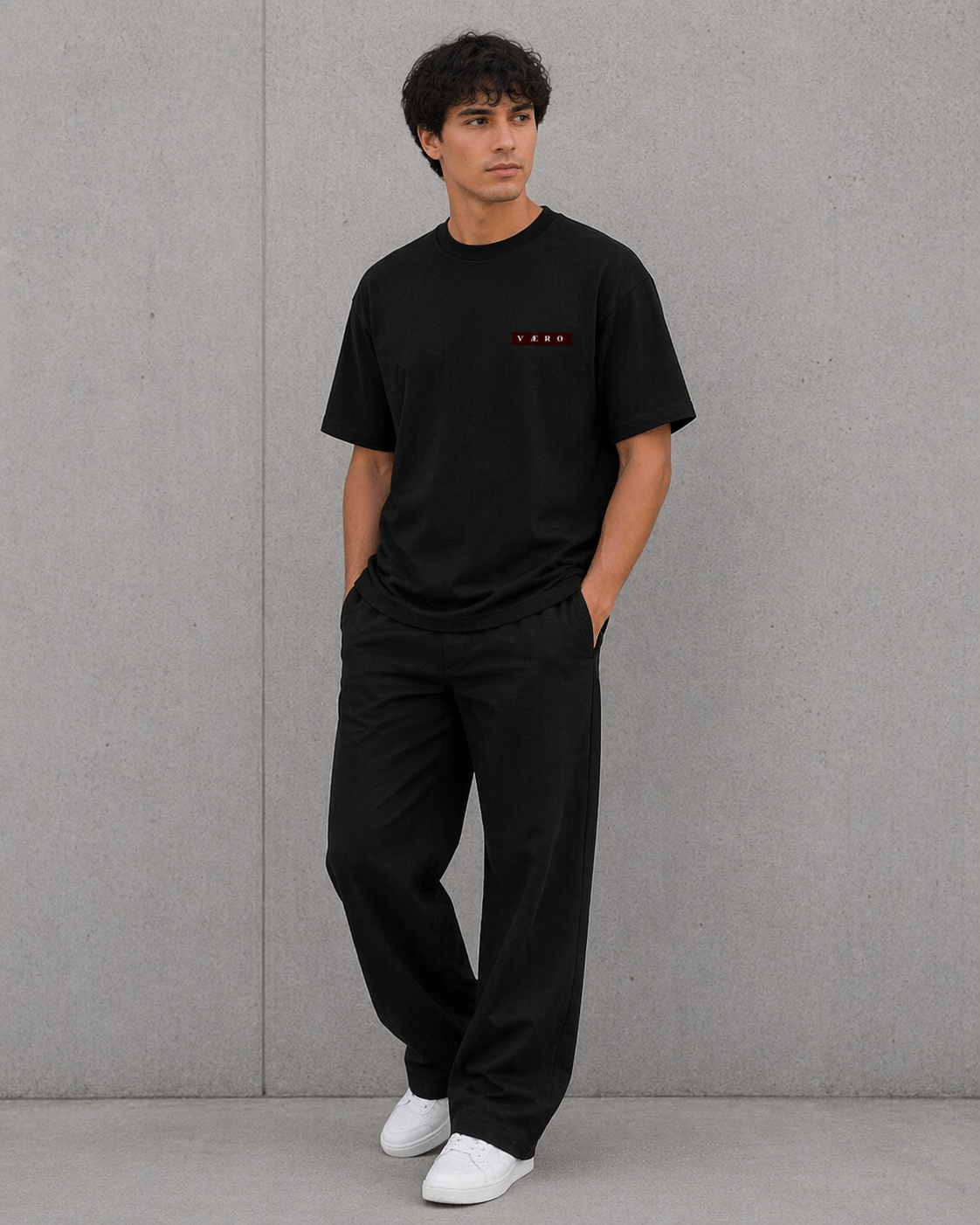 Man wearing a black t-shirt and pants standing against a gray wall.